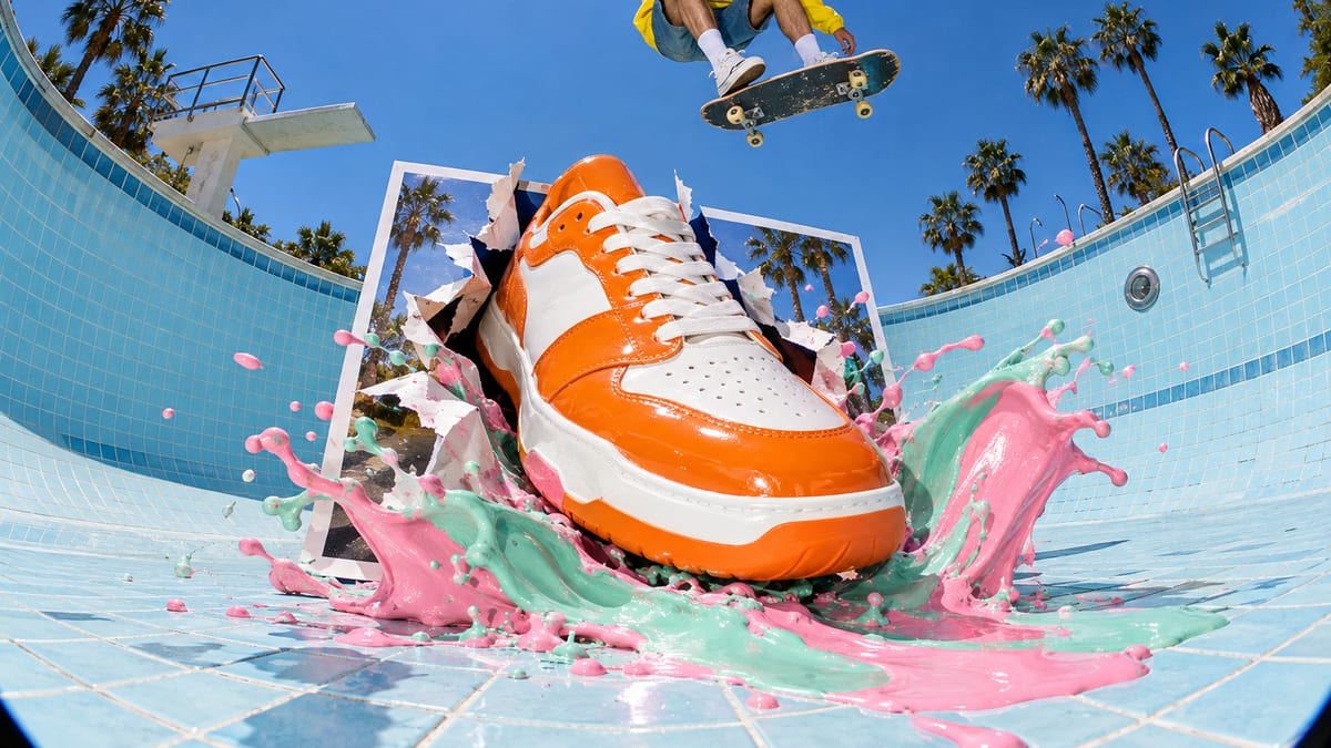 An oversized glossy sneaker bursting out of a giant paper product photo into an empty pastel swimming pool, throwing a wave of pink and mint paint while a skateboarder flies over it.