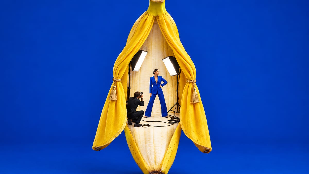 A giant peeled banana on a cobalt backdrop, its peel drawn back like theatre curtains to reveal a miniature photo shoot with a model under two softboxes.