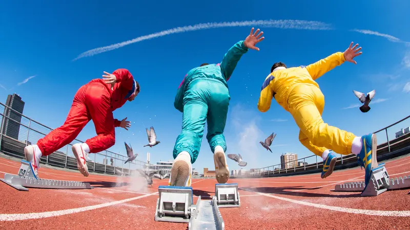 Three sprinters in red, teal and yellow retro tracksuits burst out of starting blocks on a rooftop running track